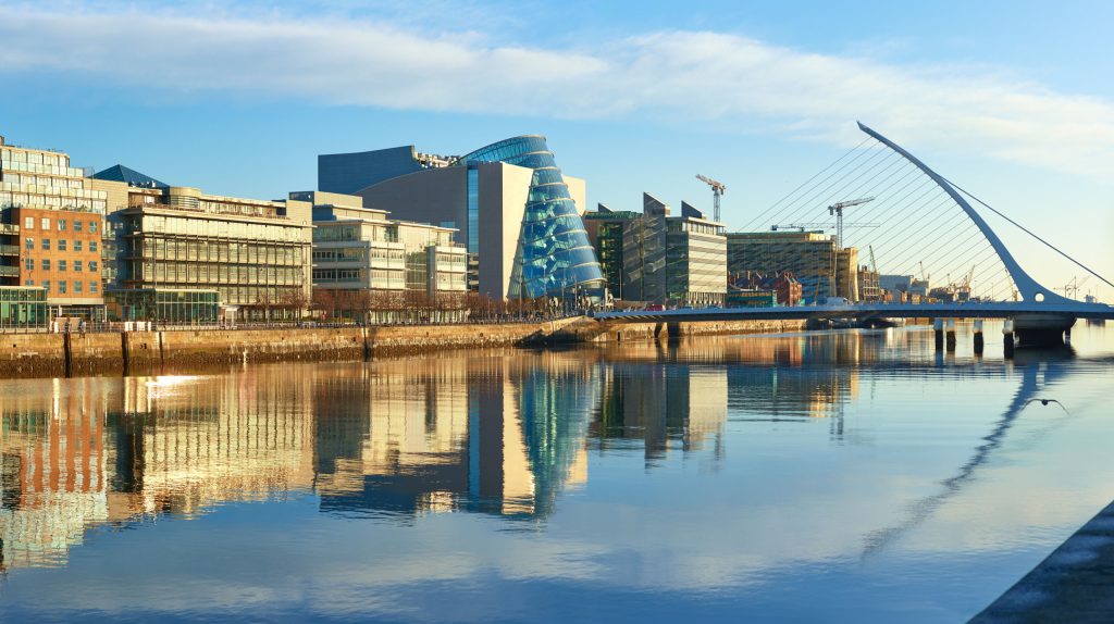 Riverfront cityscape with glass office buildings and a curved glass tower beside a calm waterway (informative image)