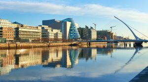 Inovacijos - strateginis verslo augimo variklis Airijos rinkoje - Verslo Klubas | Strategic Minds Society - Verslo Klubas | Strategic Minds Society Riverfront cityscape with glass office buildings and a curved glass tower beside a calm waterway (informative image)