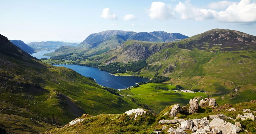 A tranquil valley with a winding blue lake surrounded by green hills and rugged mountains under a bright sky.