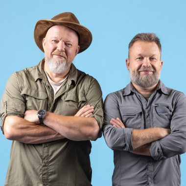 Two smiling men with gray beards standing side by side, arms crossed against a bright blue background.